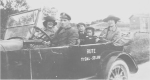 Rutekjøring med åpen personbil i 1923. Bak rattet Torstein Korsvold, og ved siden av han Aslaug Stuedal. Marit Morset sitter i baksetet med sønnene Oddmund og Tormod. Amanda og Kirsti Svelmo står ved siden av bilen.